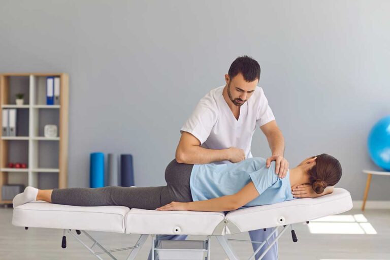 Chiropractor assessing a patient’s spine beside a massage therapist working on shoulder muscles in a clinical room.